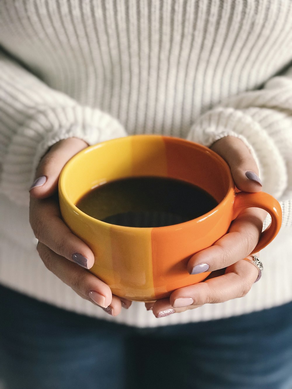Two hands in a cream sweater holding a large orange cup of coffee
